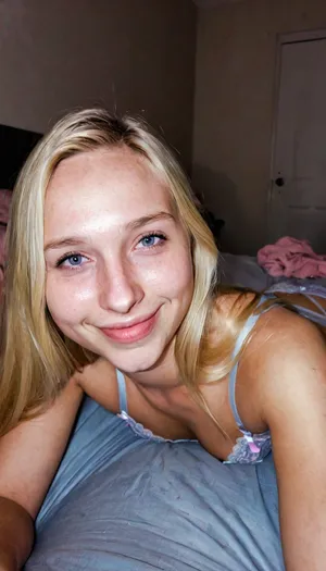 candid photo, confident 18 year old girl, lingerie, soft body, babyfat, small breasts, flat chest, long blonde hair, ((wide jaw)), ((big chin)), thin lips, big nose, eyeglasses, small body, she is laying next to you in bed, someone else in bed off-screen, dark pastel colored bedroom, cute smile, ultimate realism, poor lighting, at night, dark, night, realistic detailed skin texture, focus on realistic face, stunning eyes, skin pores, ultra realism