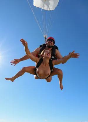 (naked woman skydiving in tandem with a naked instructor), (view from below, against a clear blue sky), (bright sunlight), (dynamic pose, falling in the air), (woman looking up, happy expression), (instructor with helmet and goggles), (woman topless, wearing safety harness), white parachute, (partially visible), (extreme sports photo), (high contrast, bright colors), professional photo, motion blur on edges, sharp focus on objects