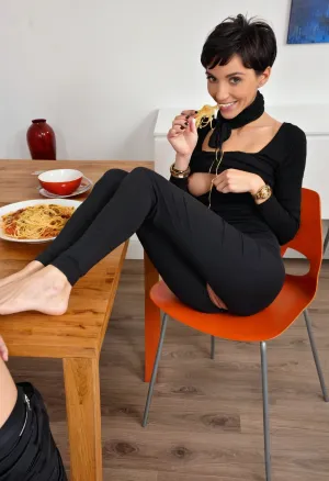 close-up photograph of a woman eating while sitting at the table with me at home with feet insider a platea of spaghetti, feet over the table, feet in platea of spaghetti, 50 years old, very thin, with a black scarf around her neck, black hair, Extreme skinny woman, messy pixie hair cut, black sweater, natural big and deflated breasts, wrinkled breast, skinny face, smile, broad shoulders, skinny ass, extremely narrow hips, skinny legs, black trousers, barefoot, black skinny pants, black nail polish, lots of steel bracelets on the right wrist and a steel watch on the left, veri long legs