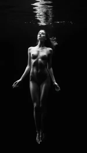 High-contrast black-and-white underwater fine art photography. A graceful female figure floats suspended in dark, still water, her body illuminated by soft, directional light from above. Her pose is elegant and fluid, evoking a sense of serenity and quiet strength, with hair drifting gently around her like ink in water. The image captures the weightless beauty of the human form beneath the surface, with subtle highlights and shadows accentuating muscle definition and curvature. The composition is minimalist, with deep blacks and gentle gradients, evoking a timeless and ethereal mood, Beautiful detailed face, (perfect eyes:1.1), (perfect skin:1.1), (perfect breasts:1.1), (perfect small pink nipples:1.1), (small areolas:1.1), (perfect vagina:1.1), (perfect body:1.1), (perfect hands:1.1), (perfect finger:1.1), (perfect fingernails:1.1), Modelshoot style, professional Photography, Photorealistic, RAW, analog, best quality, 8k, HD, DSLR, Fujifilm XT3, film grain, award winning