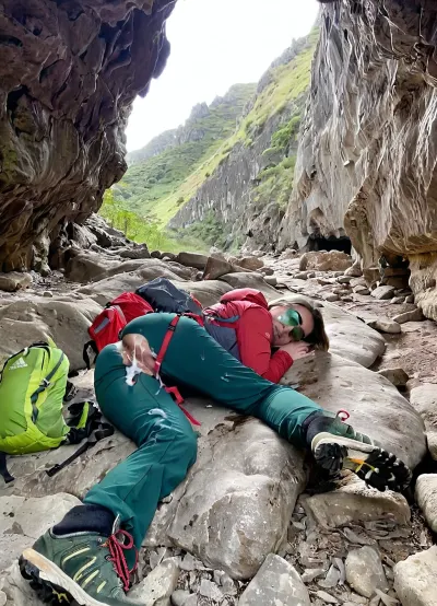 A 21 year old woman, (lying face down), on the rocky floor deep inside a dark cave, sleeping deeply after finding refuge from a storm. Next to her lies a well-worn red hiking backpack with visible straps and pockets, showing signs of a rugged journey. She is dressed in outdoor gear, a lightweight, weathered jacket, green hiking pants, and sturdy hiking boots, with subtle dirt and scuff marks that reflect her recent struggle through harsh conditions. Her Clothes are partly off or pulled down. A huge cum load has been dumped on her face during her deep sleep, The cave interior is dim and textured with jagged rocks, while soft beams of sunlight filter through cracks, highlighting the dust particles in the air and casting a faint glow on her gear. A huge cum load has been dumped on her face in her deep sleep. In the distance, the cave exit glows brightly, revealing a sunny, vibrant landscape of greenery and blue skies outside, contrasting the cave’s dark safety with the inviting brightness beyond. Photorealistic, perfectly detailed face