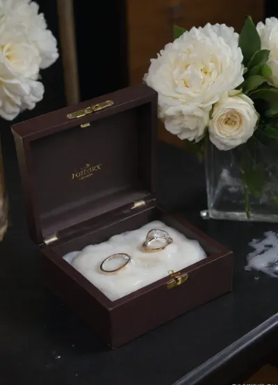 A surreal close-up of two wedding rings inside an open, elegant ring box placed on a black velvet table. Beside the box lies a delicate white bridal bouquet. Thick, glossy white slime with creamy undertones is slowly poured over the box and flowers, dripping down the sides and partially covering the rings. The lighting is soft and cinematic, enhancing the shine of the rings, the texture of the slime, and the dark contrast of the background. The scene feels poetic, artistic, and symbolically messy—blending elegance with surreal distortion.