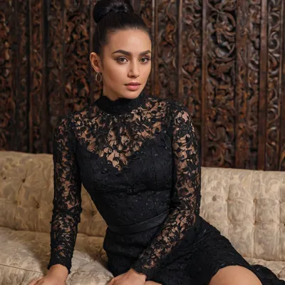 The image is a portrait of a young woman sitting on a couch. She is wearing a black lace dress with long sleeves and a high neckline. The dress is fitted at the waist and flares out at the hips. The woman has her black hair styled in a high bun and is wearing large hoop earrings. She has a serious expression on her face and is looking directly at the camera. The background is a wooden wall with a patterned wallpaper.