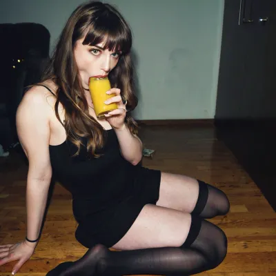 (A 22-year-old introvert brunette grunge girl with bangs and dark stockings and a short dark dress is sitting on the floor in a dark corner drinking lemonade from a glass at a party in 1997), in the background a 27-year-old tall and slim woman with long wavy dark blonde hair is visible, photo-realistic, compact camera shot, analogue film artefacts, grainy