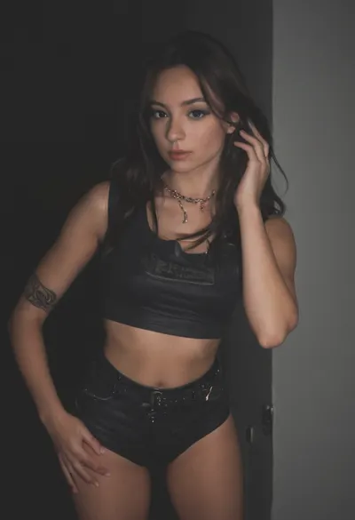 En la imagen aparecen dos personas posando en lo que parece ser una discoteca o club nocturno. La mujer, a la izquierda, viste un top de encaje negro con transparencias y tiene tetas perfectas y grandes y escote y un collar largo de cuentas moradas, junto a unos vaqueros. El hombre, a la derecha, lleva una camiseta estampada en blanco y negro con letras naranjas y sostiene un vaso en la mano derecha. Ambos miran a la cámara con gesto relajado. El entorno muestra lámparas esféricas decorativas de colores colgando del techo y focos metálicos de iluminación. Tetas