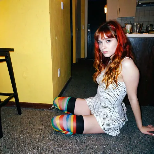 L'introverso rossa grunge ragazza con bangs e calze multicolore e un simpatico vestito di cotone corto è seduto sul pavimento in un angolo scuro bere limonata da un bicchiere a una festa nel 1997, un vecchio 27