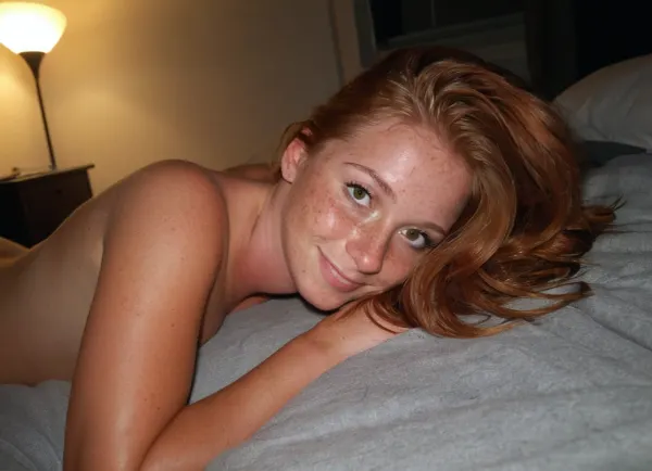 candid pov photo, stunning Amature 21 year old, stylish ginger hair, laying on her side next to you on a bed, dark dorm room, nude, small flirty smile, ultimate realism, poor lighting, at night, dark, night, realistic detailed skin texture, focus on realistic face, Sun-kissed skin, detailed realistic large pleading eyes