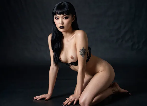 Actress Hana Mae Lee sensual figure, smooth glowing skin, brown eyes, blank semi-long hair, kneeling on a soft surface. Her back is slightly arched, hands resting on her thighs, a tattoo small at her left leg, a goth style makeup, black lipstick, black painted eyes, head tilted gently to the side with an alluring expression. Shot in a minimalist studio with dramatic lighting that creates subtle shadows across her body, ultra-detailed photorealism, perfect anatomy, lifelike detail, cinematic photography, 8K HDR..